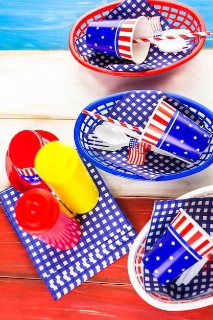 A close-up shot of a picnic setup with Fourth of July themed paper plates, cups, and napkins. Grilled burgers and hot dogs are visible, along with sides like potato salad and watermelon. The background shows a sunny park with people enjoying outdoor activities.