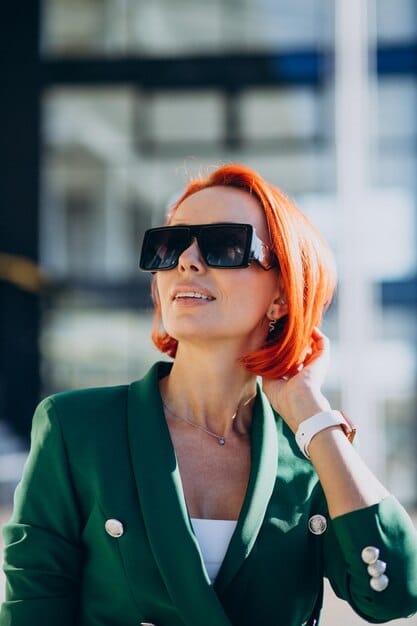 A stylish woman wearing oversized black cat-eye sunglasses and a vibrant green Shein blazer, looking confidently to the side. Her hair is sleek, and a small, elegant earring is visible.