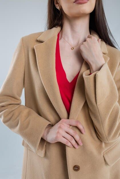 A close-up of a stylish woman's torso, featuring a well-fitted neutral-colored blazer over a crisp white blouse, with a delicate silver necklace visible. Her hands are holding a black structured handbag, conveying a sense of professionalism and elegance.