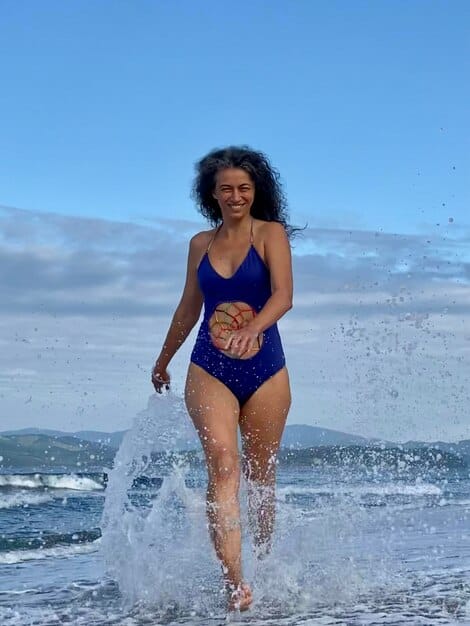 A woman with an apple body type confidently wearing a flattering Shein ruched one-piece swimsuit in a deep navy color, standing by the ocean and looking relaxed.