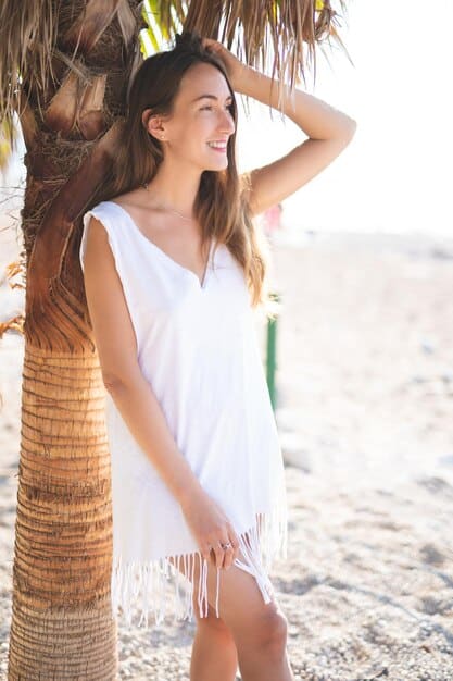 A woman stands near a beach wearing a light linen shirt over a tank top, paired with denim shorts and sandals. She is also wearing sunglasses and a wide-brimmed hat to protect herself from the sun. The overall look is relaxed and beach-appropriate, demonstrating The Art of Layering: Creating Stylish and Functional Outfits for US Weather in a warm climate.